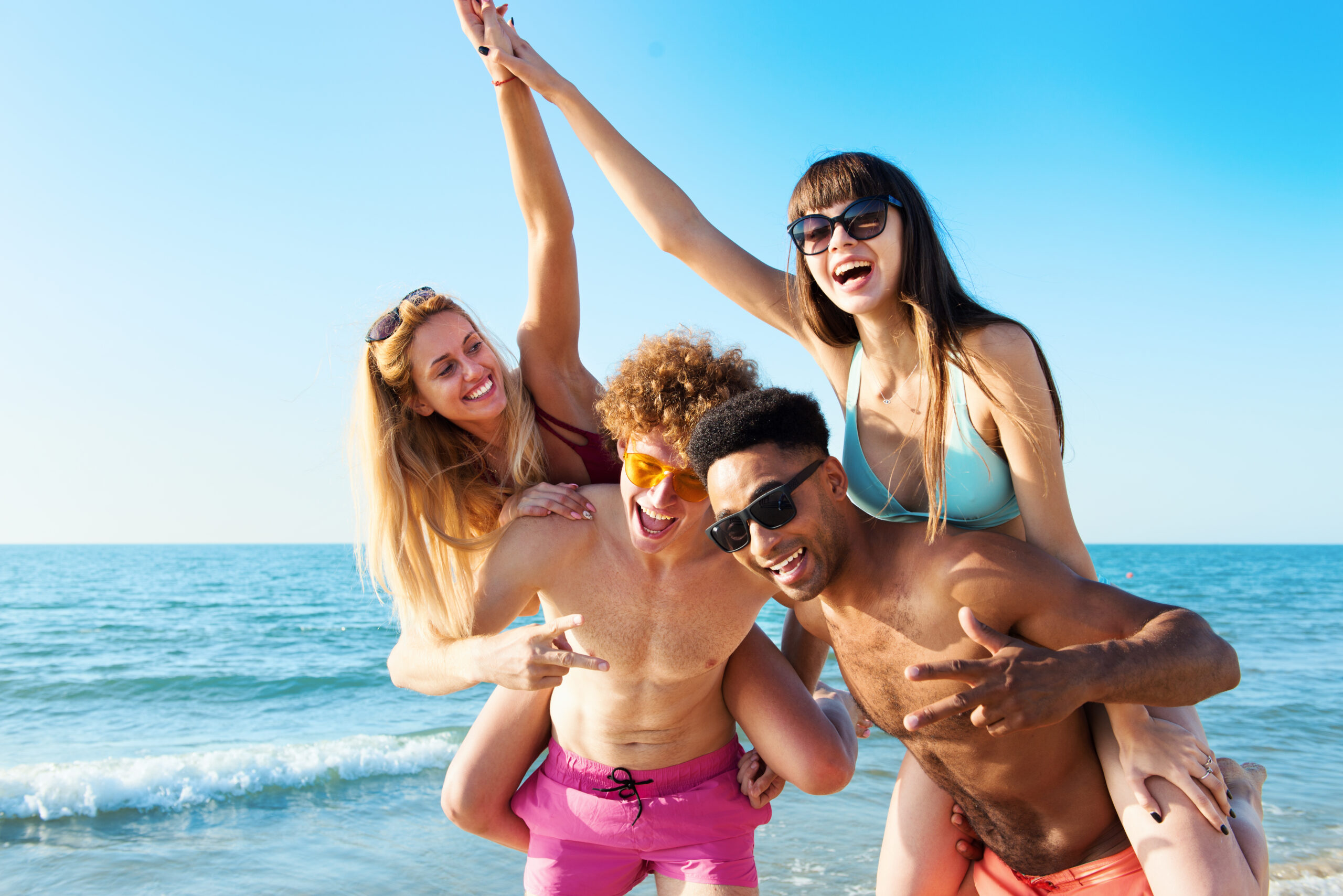 Cheerful young friends giving piggyback rides to girlfriends and enjoying summertime on the beach